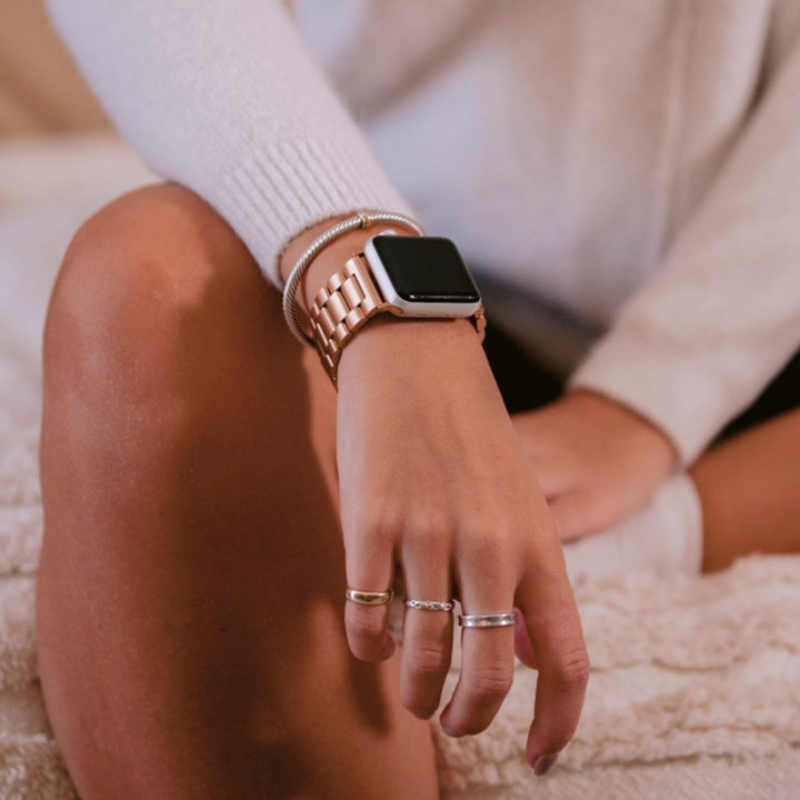 A person wearing a long-sleeve white sweater is seated with a slightly bent knee. They are adorned with multiple silver rings and a Coconut Lane Stainless Steel Apple Watch Strap in rose gold on their wrist. The background features a light-colored, textured surface, possibly a blanket or cushion.