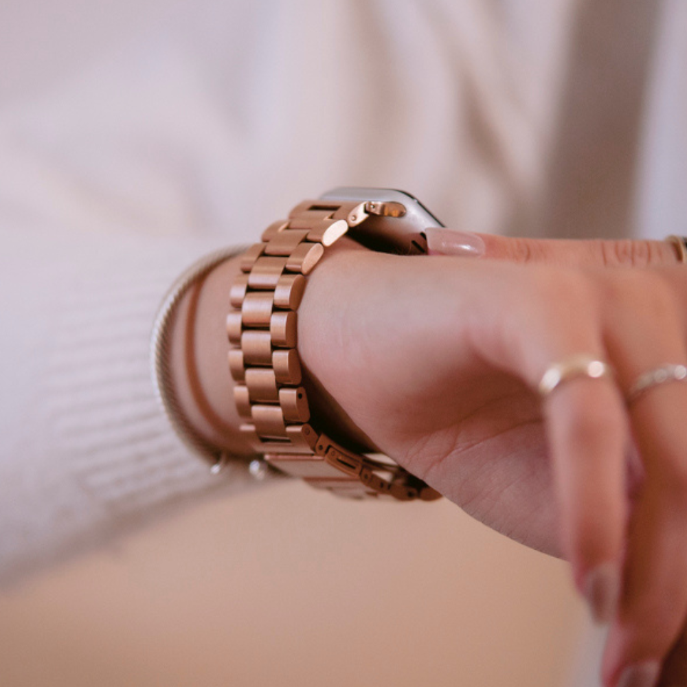 Close-up of an arm wearing a Coconut Lane Stainless Steel Apple Watch Strap in Rose Gold. The person is adjusting the watch with their other hand, while also sporting a white sweater and a silver bracelet on the same wrist. The background is soft and neutral.