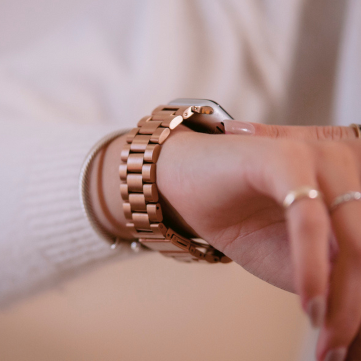 Close-up of an arm wearing a Coconut Lane Stainless Steel Apple Watch Strap in Rose Gold. The person is adjusting the watch with their other hand, while also sporting a white sweater and a silver bracelet on the same wrist. The background is soft and neutral.