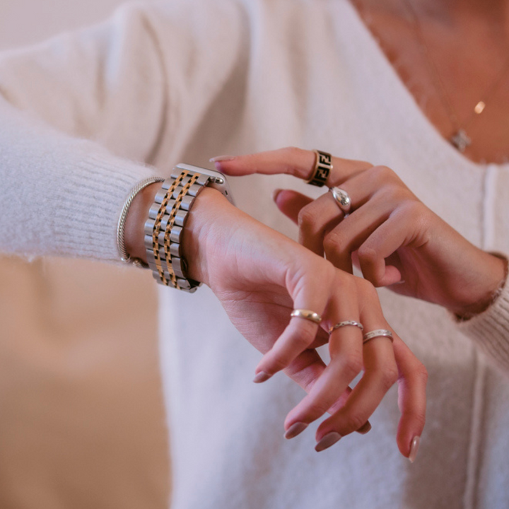 A person wearing multiple rings and a white sweater touches a stylish Apple Watch with the Coconut Lane Stainless Steel Apple Watch Strap - Silver & Gold on their left wrist.