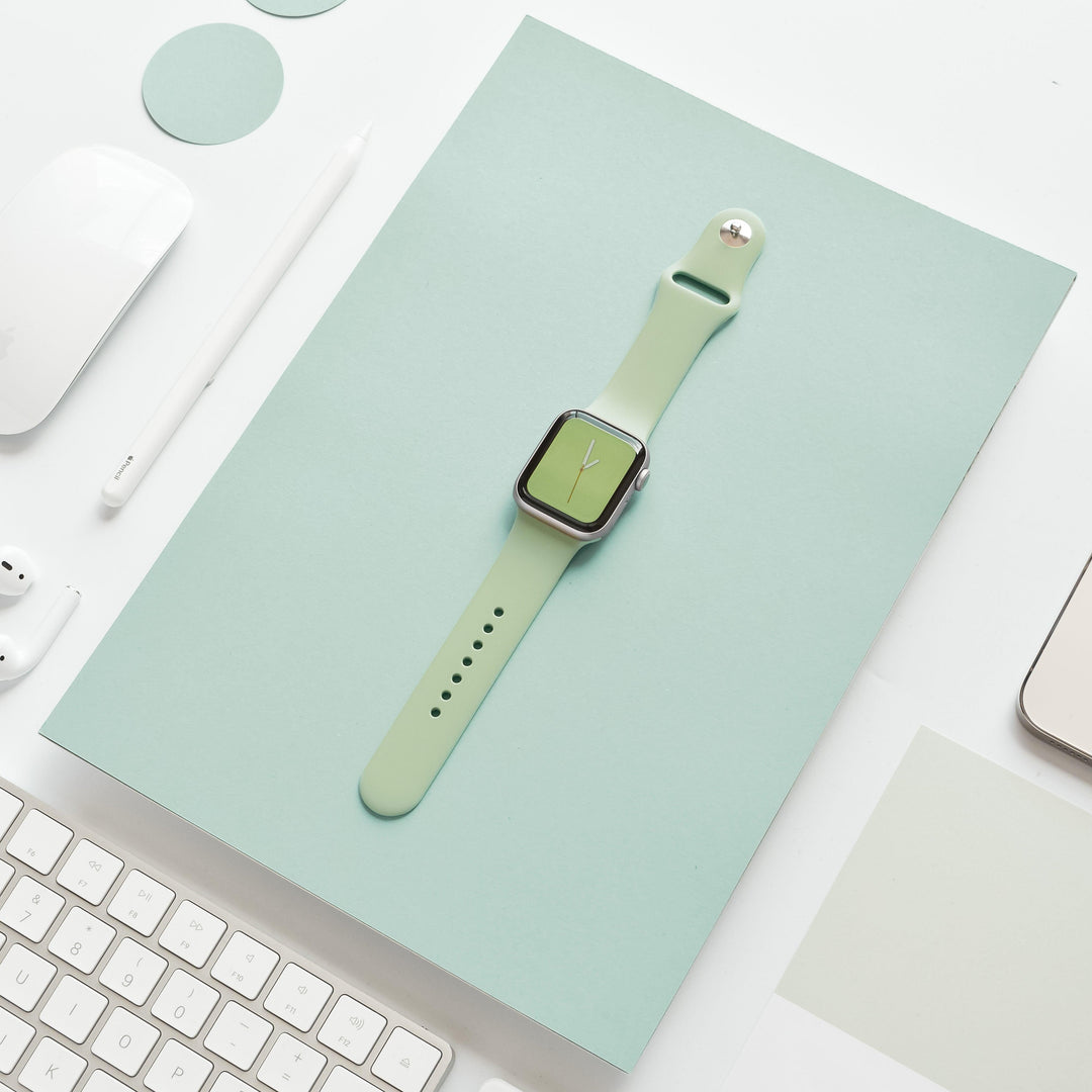 A flat lay photo showcases a smartwatch featuring the NAKD Apple Watch Strap in Sage Green from Coconut Lane, placed on matching sage-green paper. Surrounding the watch are white and light green office accessories such as a keyboard, stylus, mouse, and wireless earbuds, all set against a pristine white background.