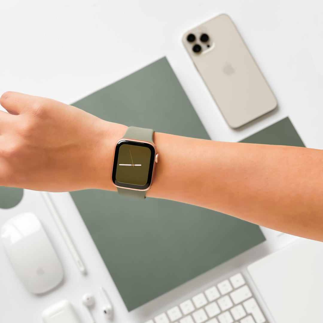 A person's wrist adorned with a smartwatch featuring a minimalist clock face and the NAKD Apple Watch Strap - Vintage Khaki by Coconut Lane, stretched over a flat lay of tech accessories. The accessories include an iPhone, keyboard, mouse, wireless earbuds, and other small objects, all boasting a sleek and modern design.