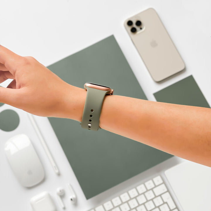 A person wearing a Coconut Lane NAKD Apple Watch Strap in Vintage Khaki is reaching over a desk. The desk features a smartphone, wireless earphones, a mouse, a mouse pad, a keyboard, and a tablet, all in coordinating shades of gray and white.