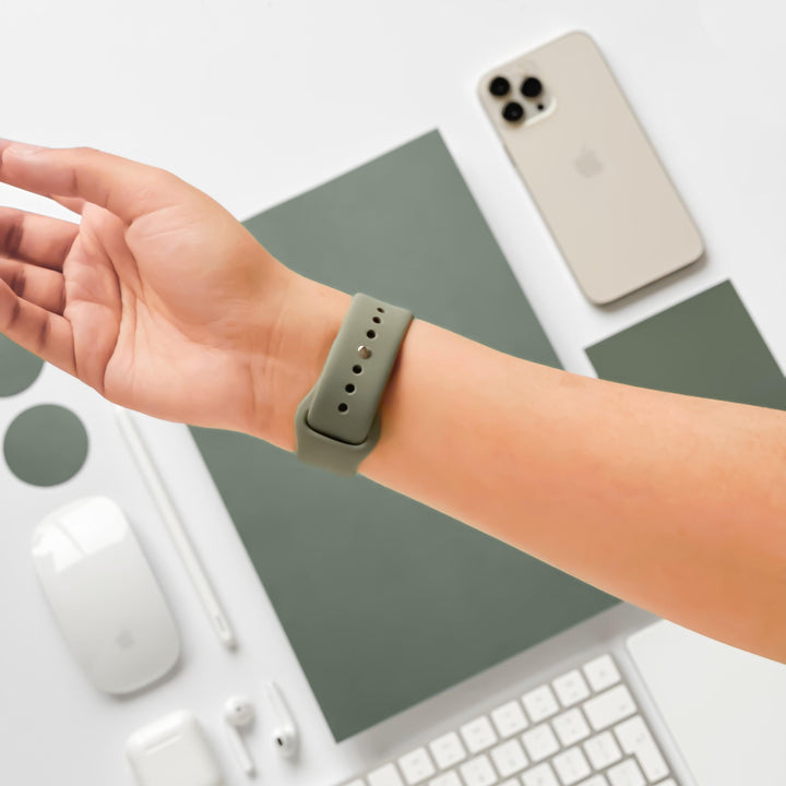 A person's wrist adorned with a NAKD Apple Watch Strap in Vintage Khaki from Coconut Lane is displayed over a desk. Nearby items include an iPhone, keyboard, wireless mouse, earphones, protective cases, and a stylus, all in shades of white and green. The background features a green notebook from the NAKD Collection.