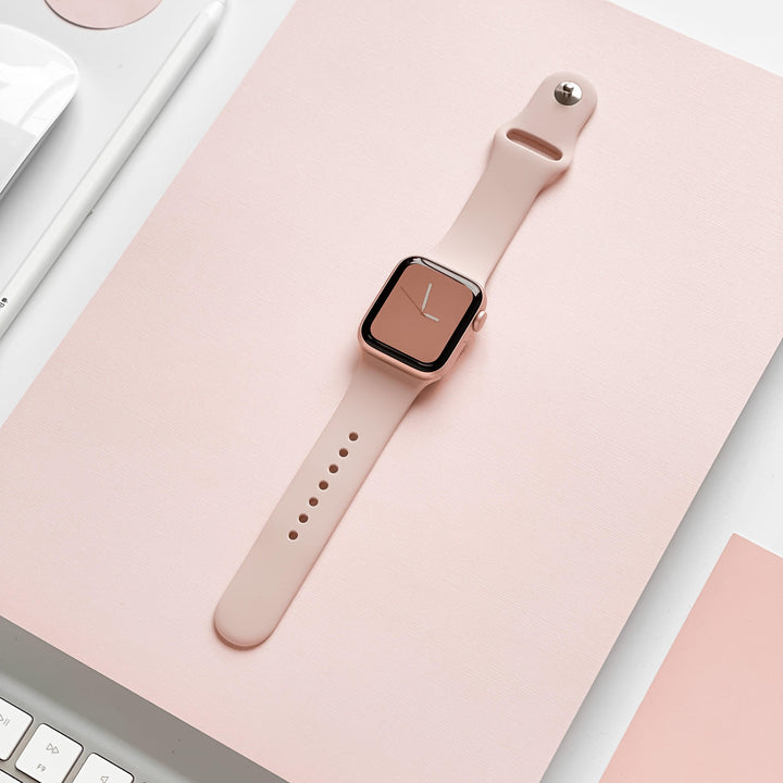 A smartwatch featuring a dusty pink silicone strap and rose gold casing is displayed on a pink surface. Nearby are parts of a white keyboard and another pink object, maintaining a cohesive pastel color scheme that is both durable and chic. The Coconut Lane NAKD Apple Watch Strap - Dusty Pink completes the stylish setup.