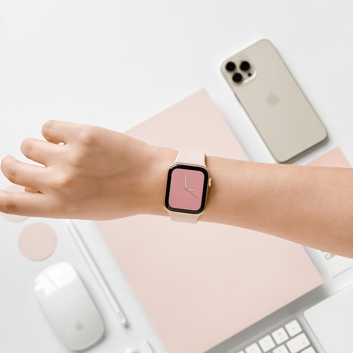 A person wearing a Coconut Lane NAKD Apple Watch Strap in Dusty Pink on their left wrist. The background features a beige smartphone, a white wireless mouse, a white key, a white pen, and a light pink notebook arranged neatly on a white surface.