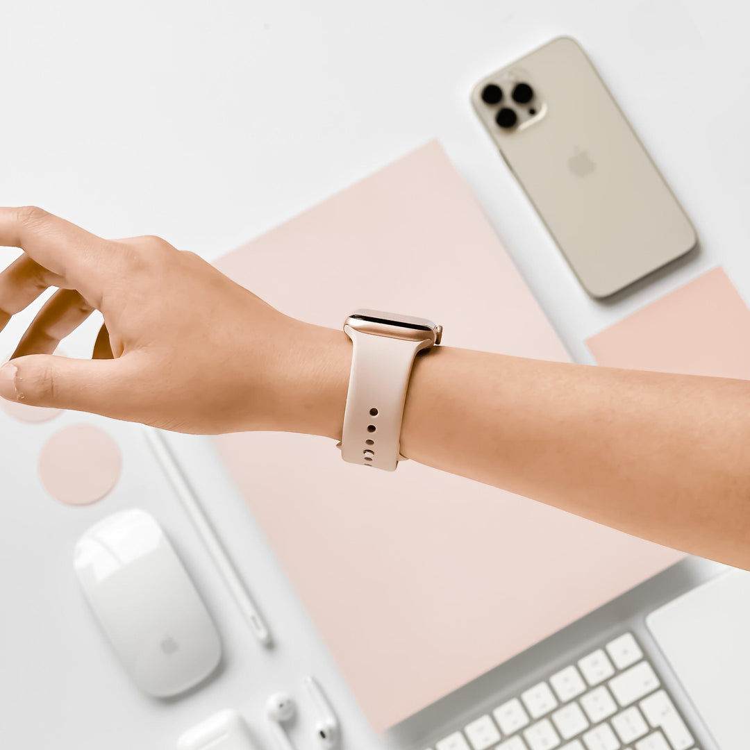 A person's left wrist wearing a smartwatch with a dusty pink NAKD Apple Watch Strap from Coconut Lane is shown in a minimalistic setup. The background includes a cream-colored smartphone, a wireless mouse, a stylus, notepads, and a keyboard, all organized neatly on a white surface.