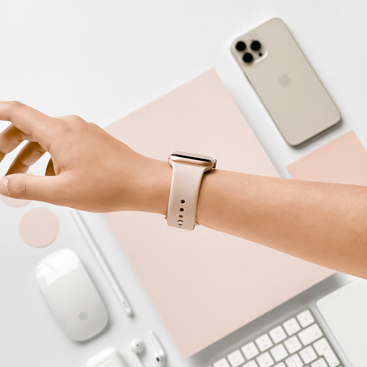 A person's left wrist wearing a smartwatch with a dusty pink NAKD Apple Watch Strap from Coconut Lane is shown in a minimalistic setup. The background includes a cream-colored smartphone, a wireless mouse, a stylus, notepads, and a keyboard, all organized neatly on a white surface.