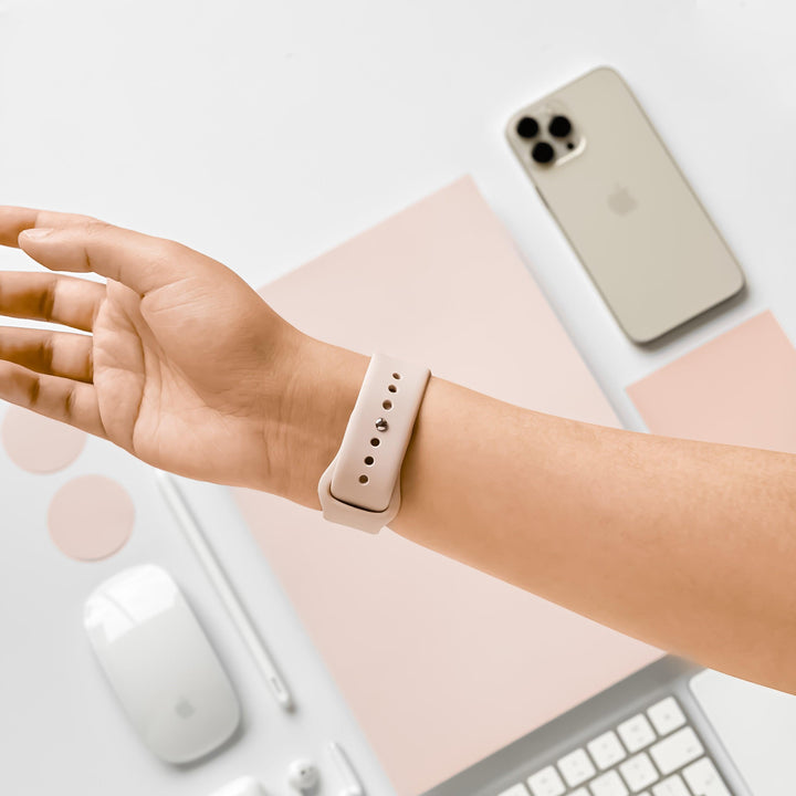 Close-up of a person's left wrist wearing the durable and chic Coconut Lane NAKD Apple Watch Strap in dusty pink silicone, with their palm facing upwards. The background features an iPhone with a matching beige case, a white computer mouse, keyboard, pen, and round stickers in a minimalistic setup.