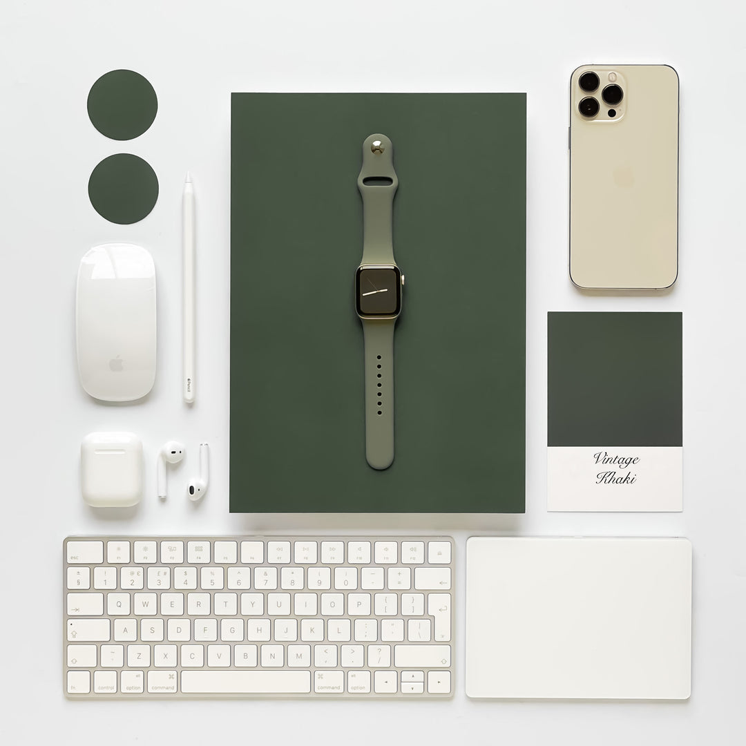 A neatly arranged flat lay of tech accessories on a white surface, featuring an Apple Watch with a NAKD Apple Watch Strap - Vintage Khaki by Coconut Lane, iPhone, wireless earphones, charger, keyboard, mouse, stylus, and trackpad. All items are coordinated with a green color theme and two circular green swatches.