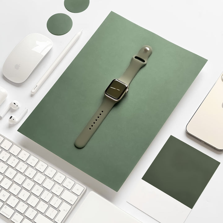 A neatly arranged desk features a green smartwatch with the NAKD Apple Watch Strap - Vintage Khaki by Coconut Lane placed on a matching green folder. Surrounding it are a white keyboard, a computer mouse, a stylus, AirPods, a smartphone, and two green circular objects. A photo print lies to the right of the watch.