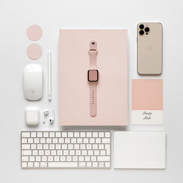 Neatly arranged tech accessories in soft pink and white tones, including a smartwatch with the durable and chic NAKD Apple Watch Strap - Dusty Pink by Coconut Lane, smartphone, keyboard, mouse, pencil, earbuds, charging cases, color swatches, and a notebook. The setup exudes a minimalist and organized aesthetic.