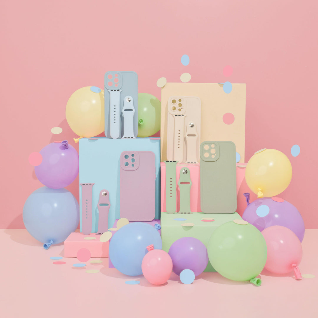 A vibrant and playful display features a colorful arrangement of pastel-colored phone cases and smartwatches, including the sleek NAKD Apple Watch Strap in Sage Green by Coconut Lane. The background is light pink with balloons in various pastel shades and scattered confetti, perfectly showcasing the accessories.