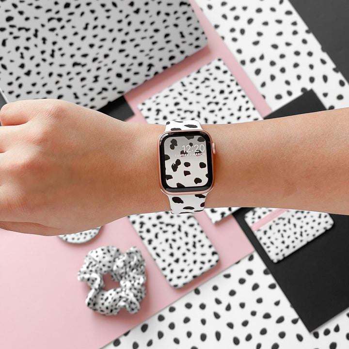 A person's wrist wears a Coconut Lane smartwatch with the Monochrome Spots Apple Watch Strap featuring a white and black Dalmatian print. The watch screen mirrors the Dalmatian print background. Various items from Coconut Lane's Monochrome Spots Collection, including notebooks and a scrunchie, are arranged on a pink and white surface.