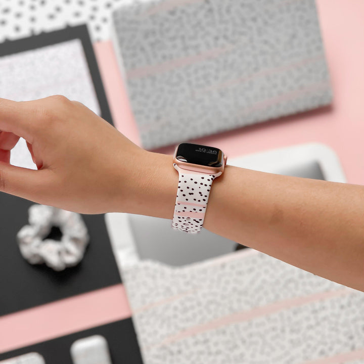 A person sporting a Coconut Lane Pink Dalmatian Apple Watch Strap adorned with black speckles on their wrist. The backdrop showcases a meticulously organized workspace featuring an array of items like a scrunchie, notebook, and assorted stationery, all flaunting matching patterns.