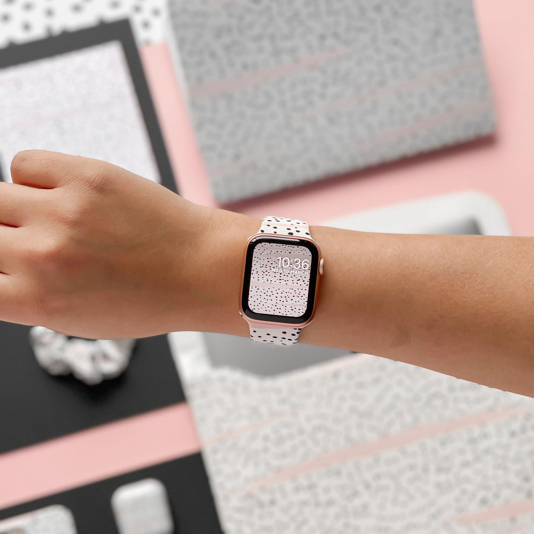 Close-up of a person wearing an Apple Watch with a Coconut Lane Pink Dalmatian Apple Watch Strap. The watch face shows 10:36, reflecting the unique speckled pattern of the strap. In the background, various items feature matching speckled designs on a pink and gray surface.