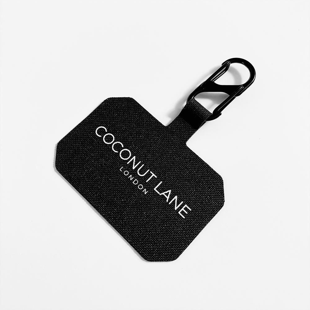 A stylish phone accessory, the Universal Phone Patch from Coconut Lane, features a black fabric luggage tag with "COCONUT LANE LONDON" in white text. It is attached to a black plastic carabiner clip, all set against a plain white background for secure phone carrying.