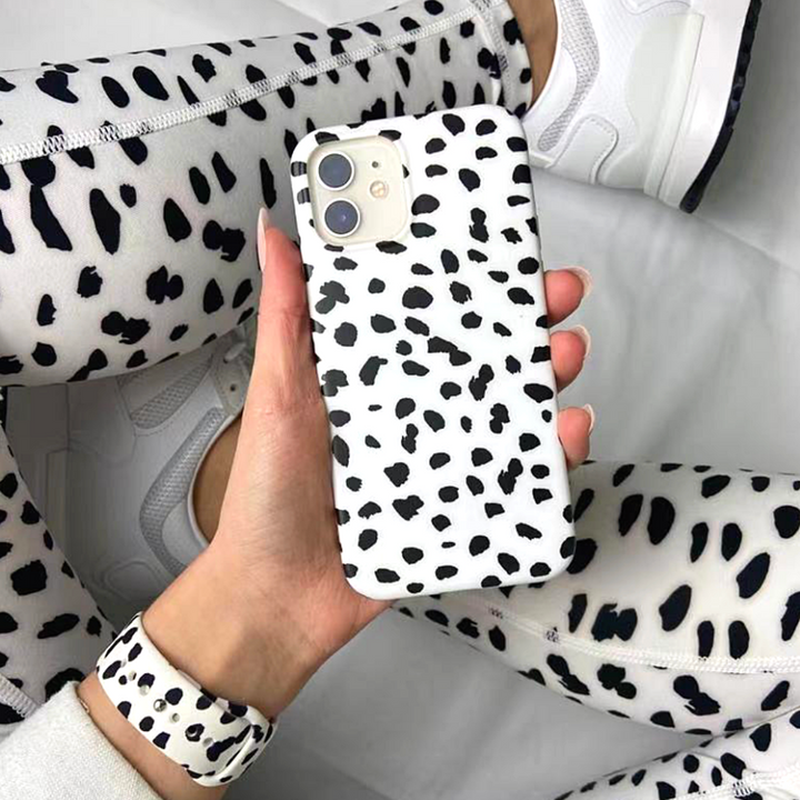 A person wearing white sneakers and monochrome spotted leggings, plus a Coconut Lane Monochrome Spots Apple Watch Strap, holds an iPhone adorned with a matching silicone case. The entire look is set against a pristine white background.