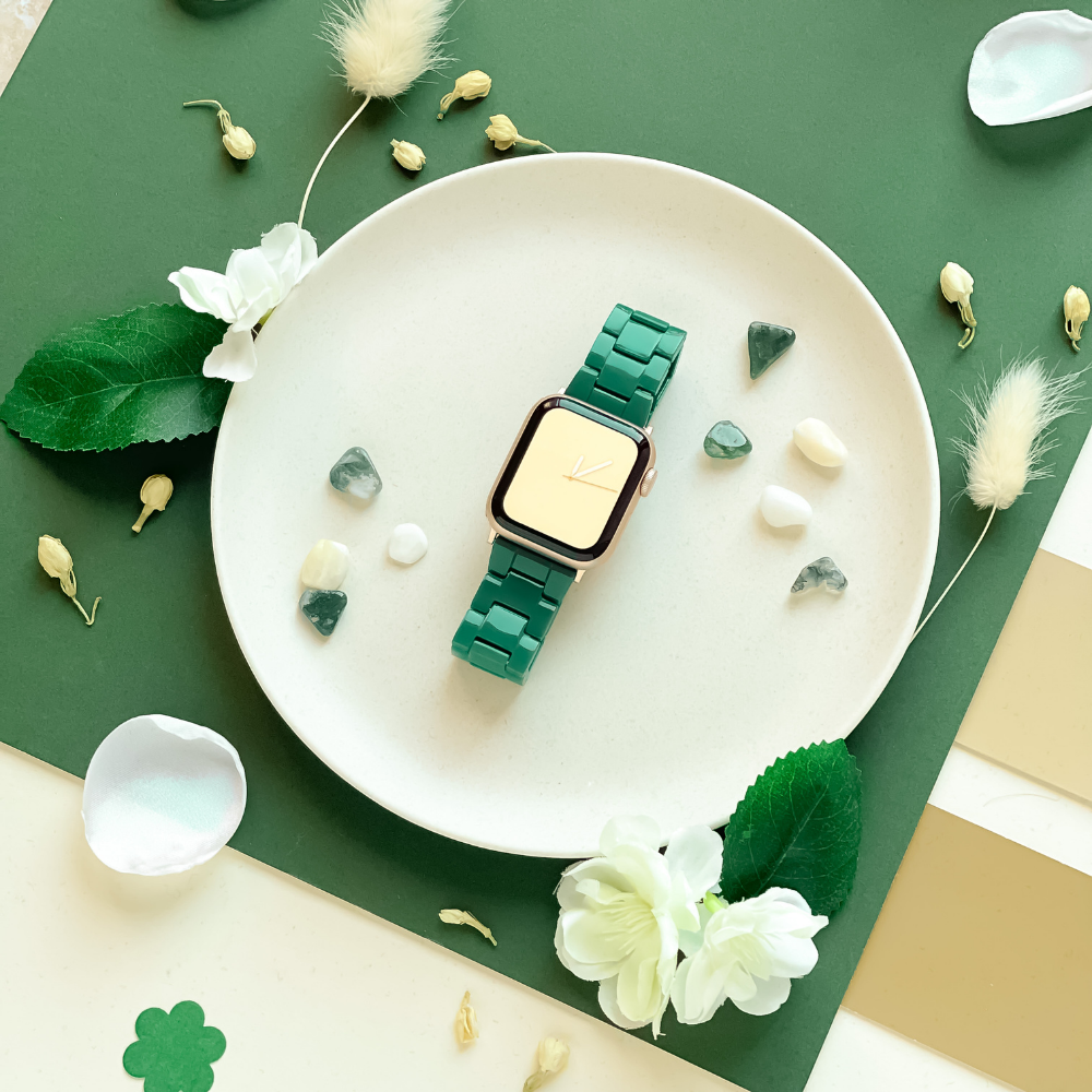 A green wristwatch with a gold square face is placed on a white plate. Surrounding the plate are green and white decorations, including leaves, small white flowers, and stones. The background is an arrangement of green, white, and light beige paper—complemented elegantly by the Luxe Green Apple Watch Strap from Coconut Lane.