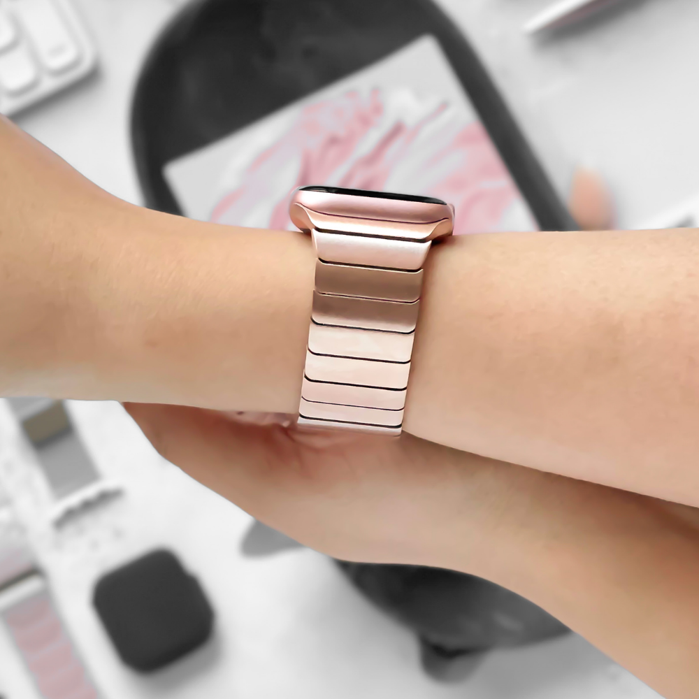 A person is wearing a rose gold smartwatch on their wrist, featuring a Slinky Apple Watch Strap - Rose Gold by Coconut Lane with a matching metal link band. The background includes various blurred objects such as another watch band, possibly for different Apple Watch models, a smartphone, and other indistinct items.