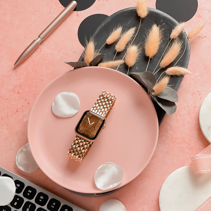 A gold smartwatch with a brown face lies on a pink plate with white flower petals, featuring the Mermaid Watch Strap - Rose Gold by Coconut Lane. Behind it, beige dried flowers and dark green leaves are arranged on a black dish. A silver pen and part of a keyboard are visible on the pink background.