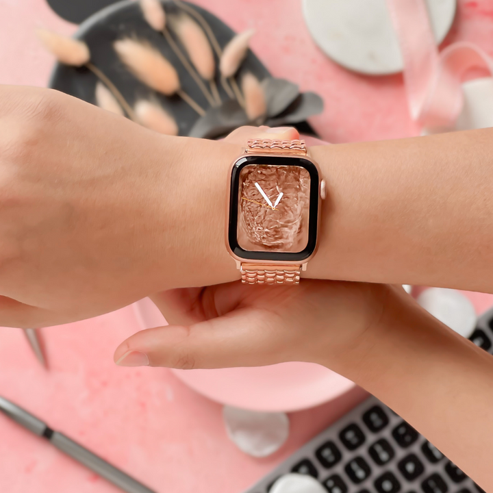 Close-up of a person's wrist wearing a smartwatch with the Coconut Lane Mermaid Watch Strap in Rose Gold. The watch face displays an abstract, textured design with a minimalist 'V' symbol. The background includes a keyboard, pen, ribbon, and tray of dried floral decorations against a pink surface.