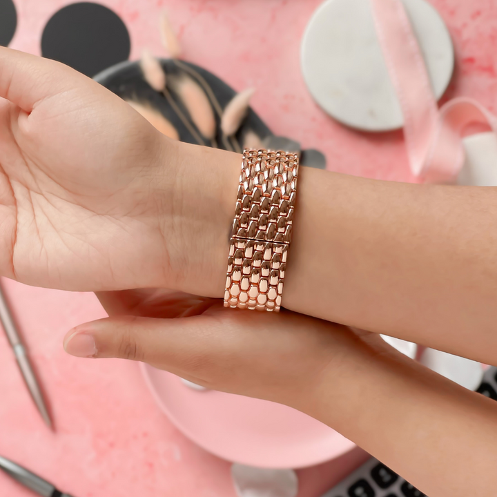 A hand with neatly manicured nails holds a wrist adorned with Coconut Lane's Mermaid Watch Strap in Rose Gold. The background is soft pink, featuring visible items such as makeup brushes, ribbon, a pen, and a keyboard.