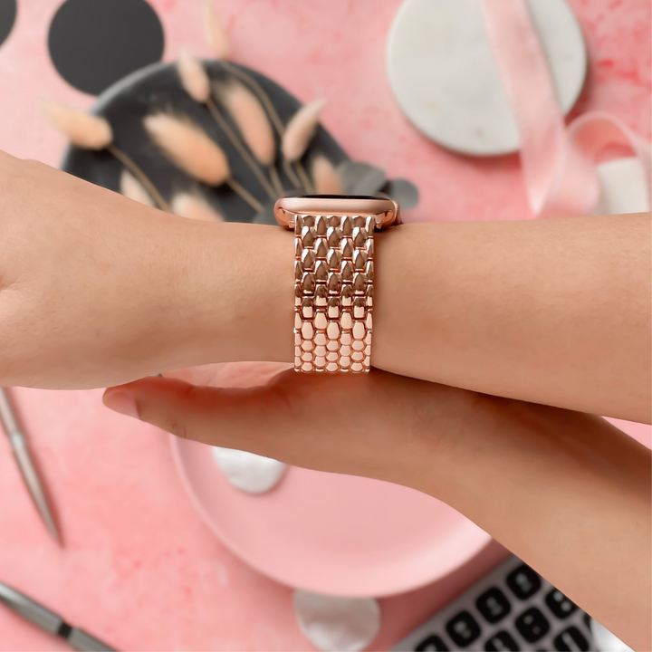 A person displays a wrist adorned with Coconut Lane’s Mermaid Watch Strap in Rose Gold. The background is pink and features various makeup brushes, a ribbon, a pen, and a keyboard, creating a stylish and feminine setting. A free link remover tool is included for easy adjustments.