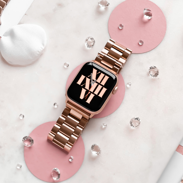 A rose gold smartwatch with a Coconut Lane Stainless Steel Apple Watch Strap rests on a pink and white marble surface adorned with pink circles and scattered clear crystals. The watch face displays a playful arrangement of Roman numerals. The background includes soft fabric elements nearby and small water droplets.