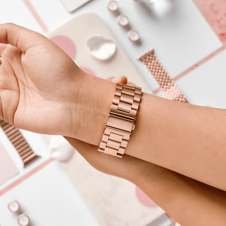 Close-up of a person putting on a Coconut Lane Stainless Steel Apple Watch Strap in Rose Gold. In the background, various accessories and other watch bands, including adjustable stainless steel metal straps for different Apple Watch models, are scattered on a light-colored surface.