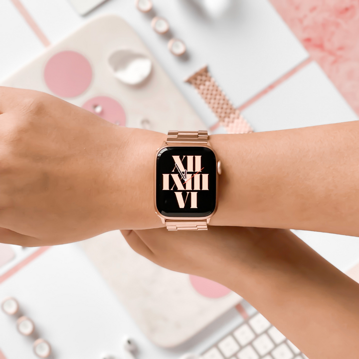 A person's wrist showcasing a smartwatch featuring a round gold frame and a pink band. The screen displays large white Roman numerals. The background includes a keyboard, another watch band, and various accessories in pastel colors, beautifully highlighting the elegance of the Stainless Steel Apple Watch Strap - Rose Gold by Coconut Lane.