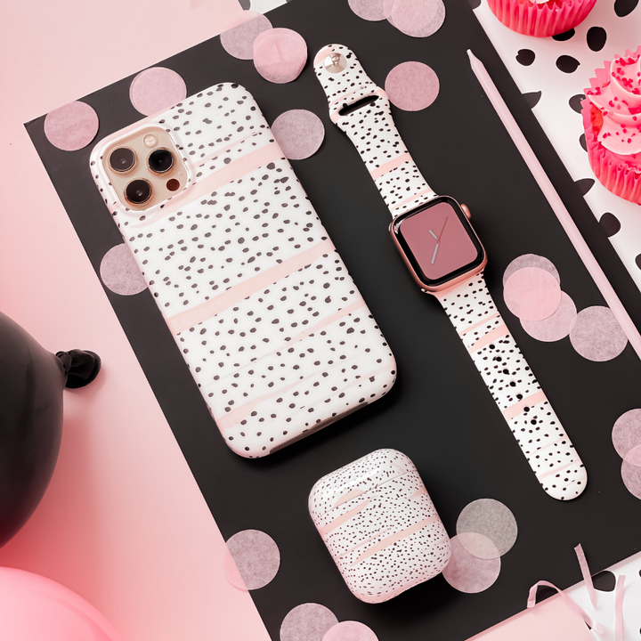 A flat lay image features a smartphone, smartwatch, and earbuds case all with matching white and black speckled patterns on a black and pink polka-dotted background. Decorative pink cupcakes and balloons are visible in the background, complementing the Coconut Lane Pink Dalmatian Apple Watch Strap perfectly.