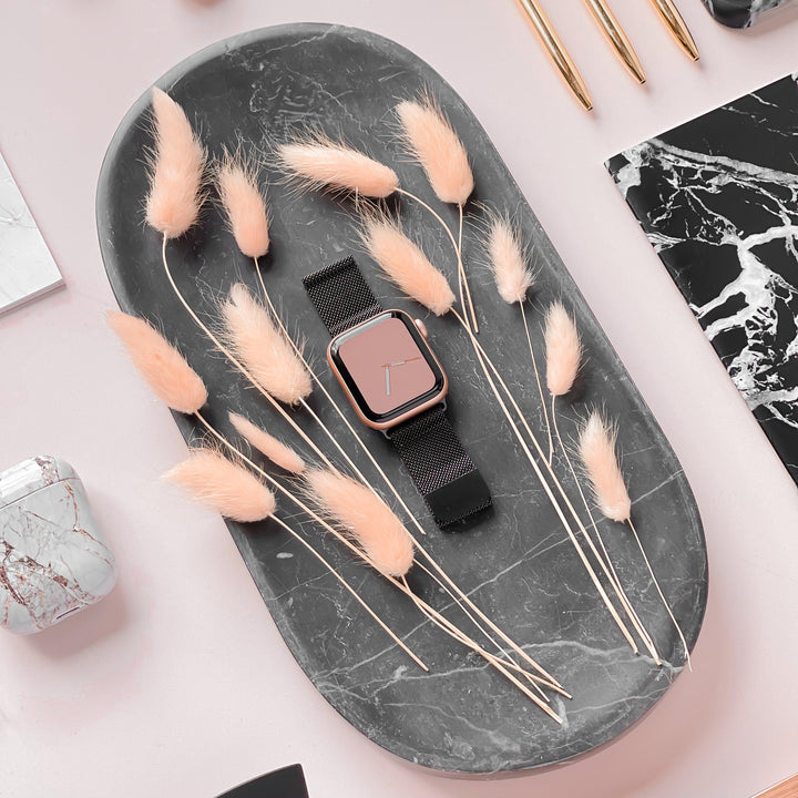 A rectangular black marble tray showcases an Apple Watch with a rose gold face and a Coconut Lane Magnetic Chain Apple Watch Strap in black, accented by pale peach-colored dried bunny tail grasses. A pink surface in the background features a marble notebook, pens, and a marble-patterned phone case. The sleek stainless steel clasp of the Apple Watch strap adds an extra touch of elegance.