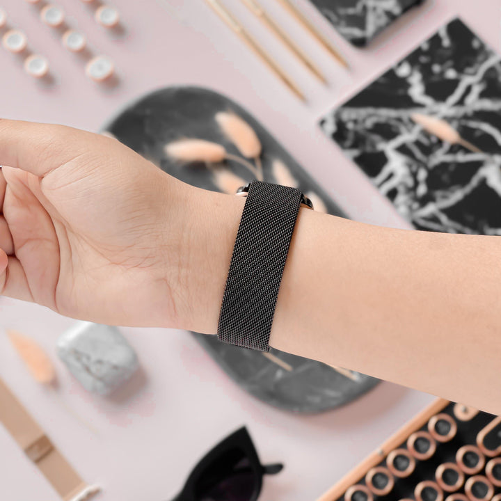 A person's hand is adorned with the Magnetic Chain Apple Watch Strap - Black by Coconut Lane, showcasing its sleek, modern design complete with a stainless steel clasp. The background features a flat lay arrangement of accessories such as sunglasses, a marble tray, a marbled notebook, and various small items in neutral tones.