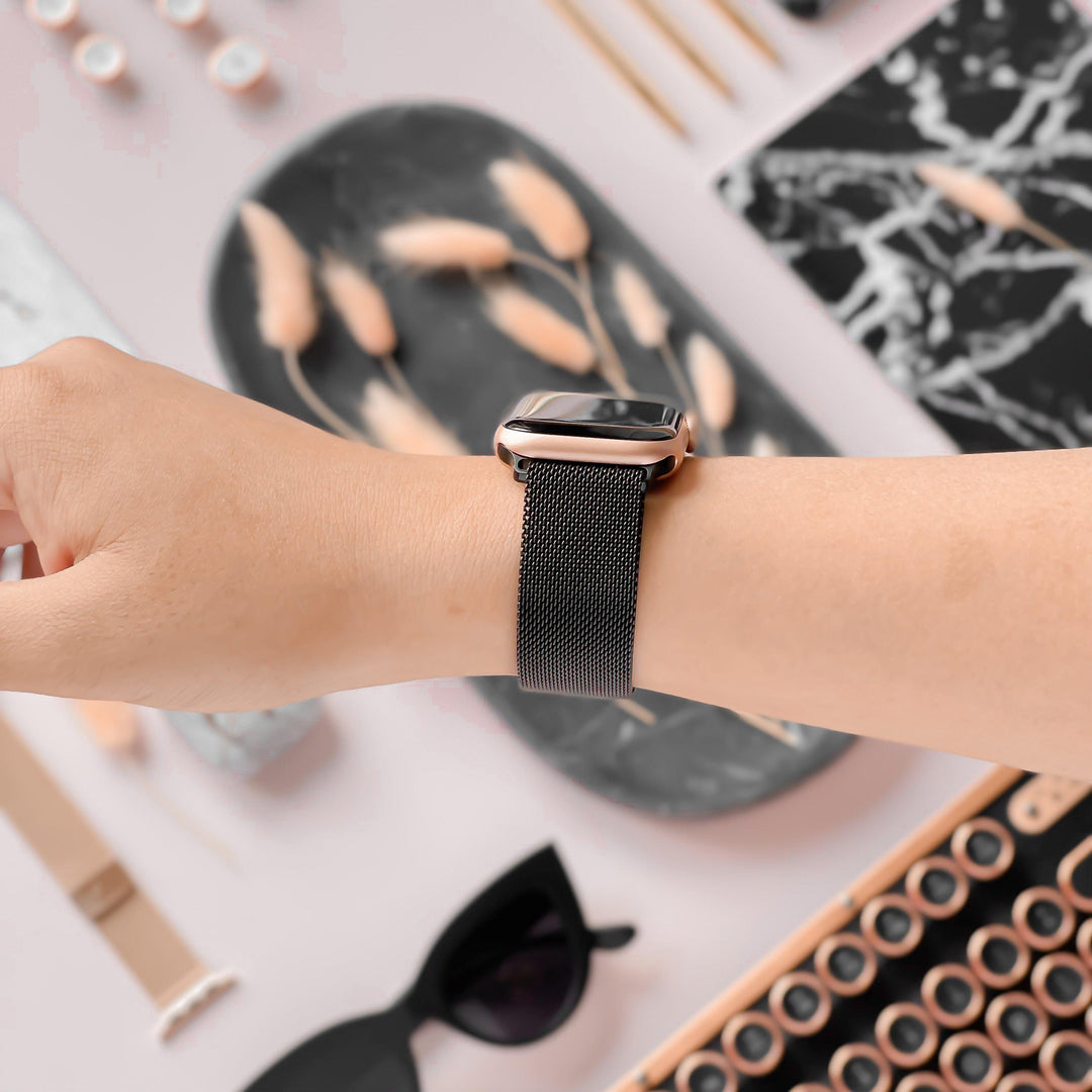A person’s wrist adorned with the Magnetic Chain Apple Watch Strap in Black by Coconut Lane, featuring a sleek metallic mesh band and a stainless steel clasp. The background showcases an array of stylish items, including sunglasses, a black marble-patterned notepad, and decorative objects tastefully arranged on a light surface.