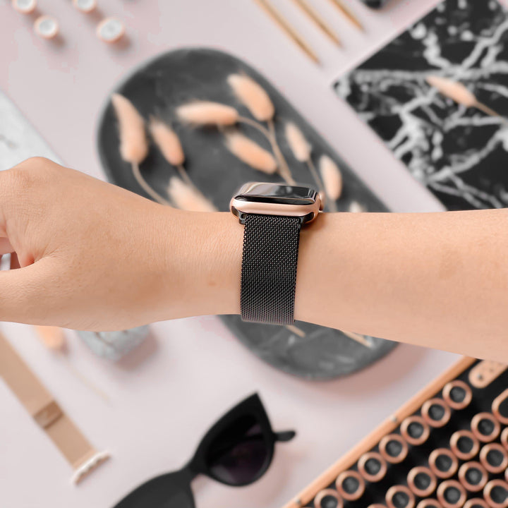 A person’s wrist adorned with the Magnetic Chain Apple Watch Strap in Black by Coconut Lane, featuring a sleek metallic mesh band and a stainless steel clasp. The background showcases an array of stylish items, including sunglasses, a black marble-patterned notepad, and decorative objects tastefully arranged on a light surface.