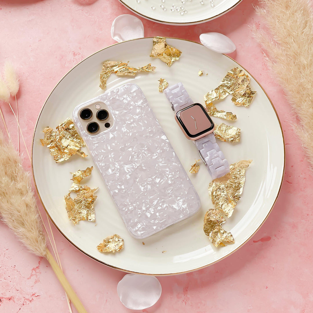 A pastel pink-themed flatlay showcases an iPhone enclosed in a textured white case and a white wristwatch fitted with Coconut Lane's Luxe Pearl Apple Watch Strap, elegantly placed on a decorative plate embellished with gold leaf details. The soft background is enhanced by pink petals and fluffy beige pampas grass.